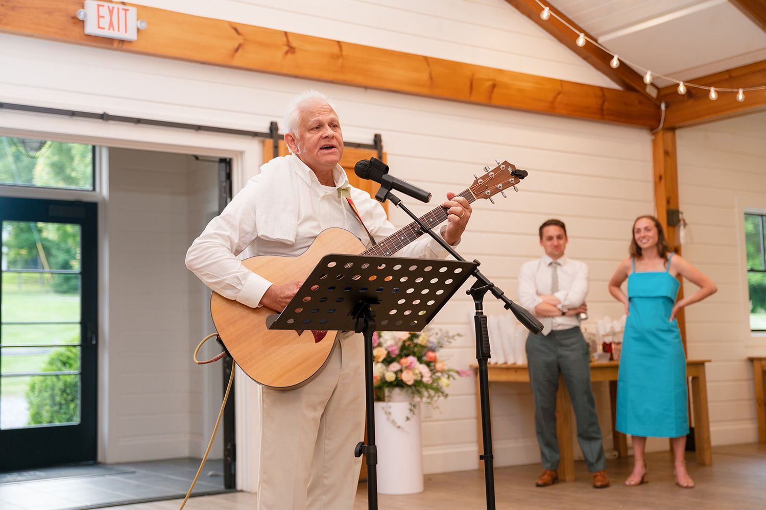 A musician performs with an acoustic guitar at a music stand while two people watch from the side in a bright venue.