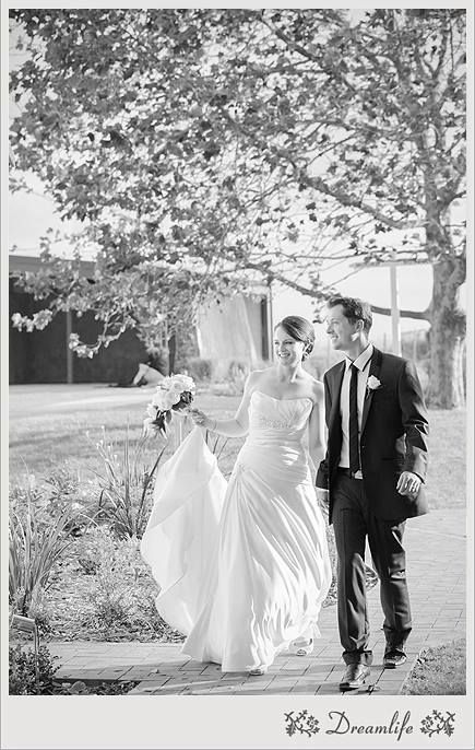 A couple walks together along a garden path under blooming spring trees in an elegant black and white wedding photo.