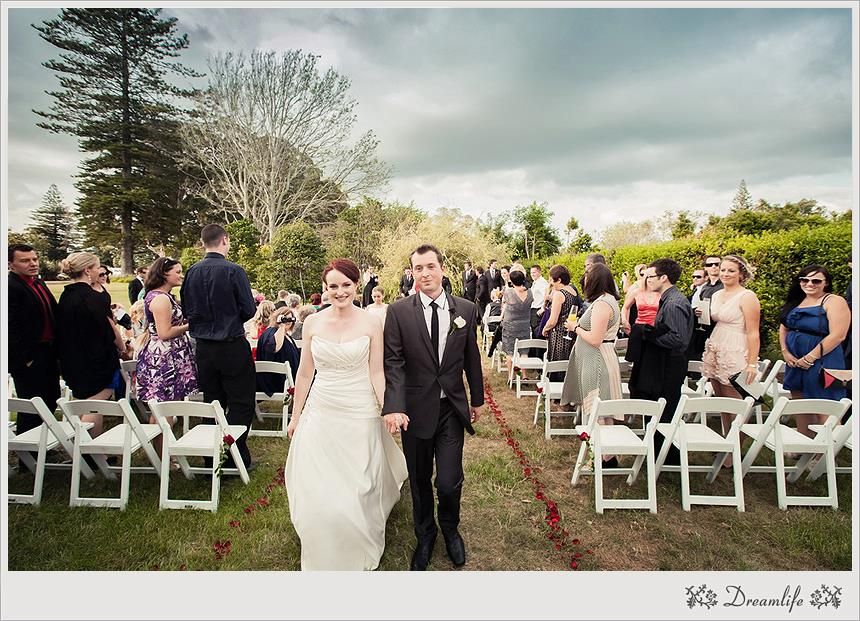 A scenic outdoor wedding ceremony at a garden venue with white chairs and guests standing along a path surrounded by trees.
