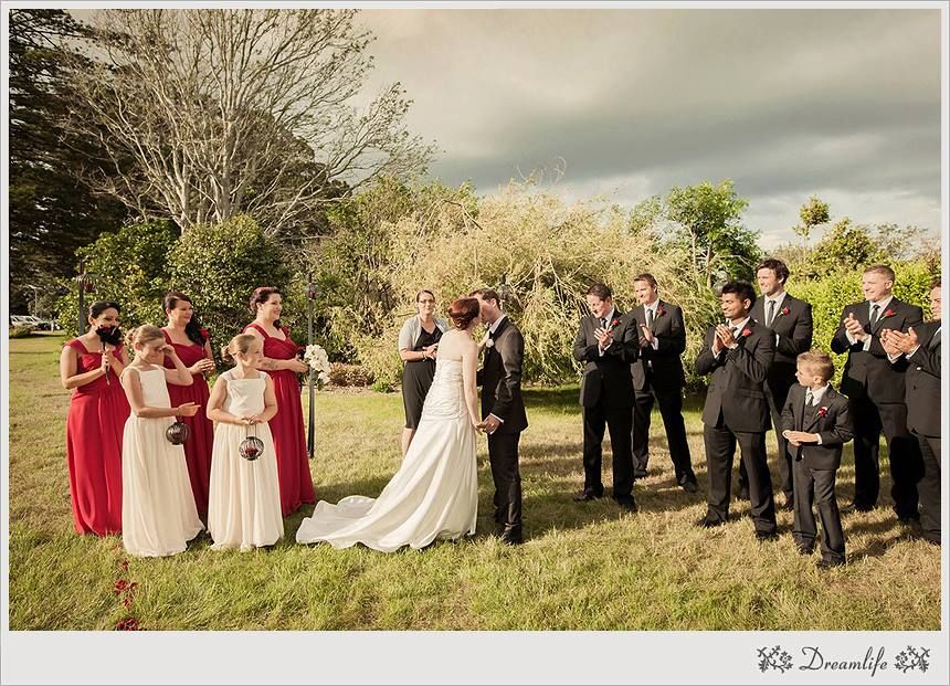 A wedding party in red bridesmaid dresses and black suits stands on a grassy hill under cloudy skies during an outdoor ceremony.