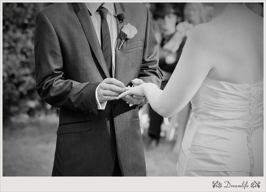 Black and white wedding ceremony photo showing couple exchanging rings, with groom in suit and bride in white dress.