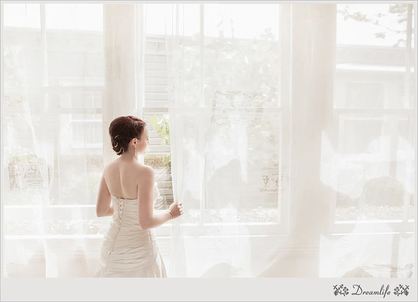 Person in white wedding dress stands by window with flowing sheer curtains in bright sunlit room.