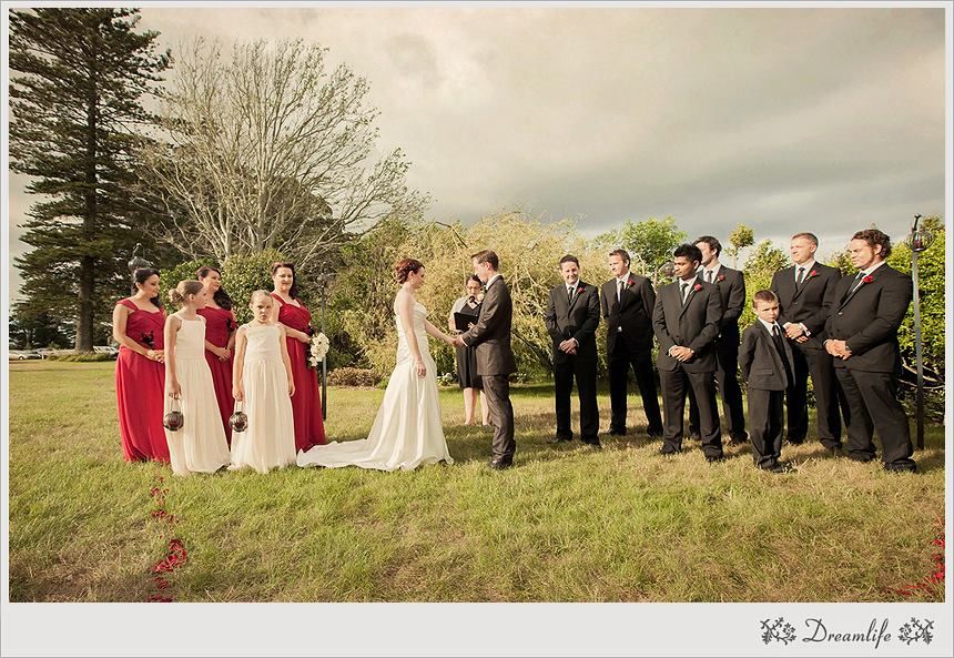 Wedding party members stand in a row along a grassy hillside during an outdoor ceremony in varied formations.
