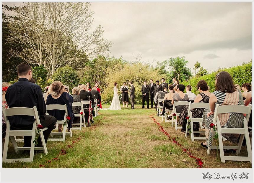 Outdoor wedding ceremony under open sky with white chairs set up in rows on a grassy path lined by trees.