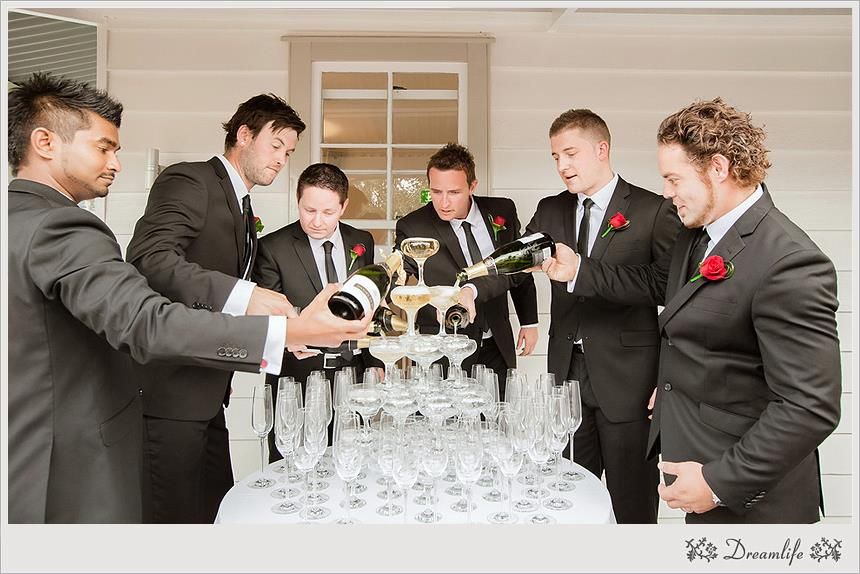 Wedding party gathers around champagne tower as groomsmen in black tuxedos carefully pour bubbly into crystal glasses.