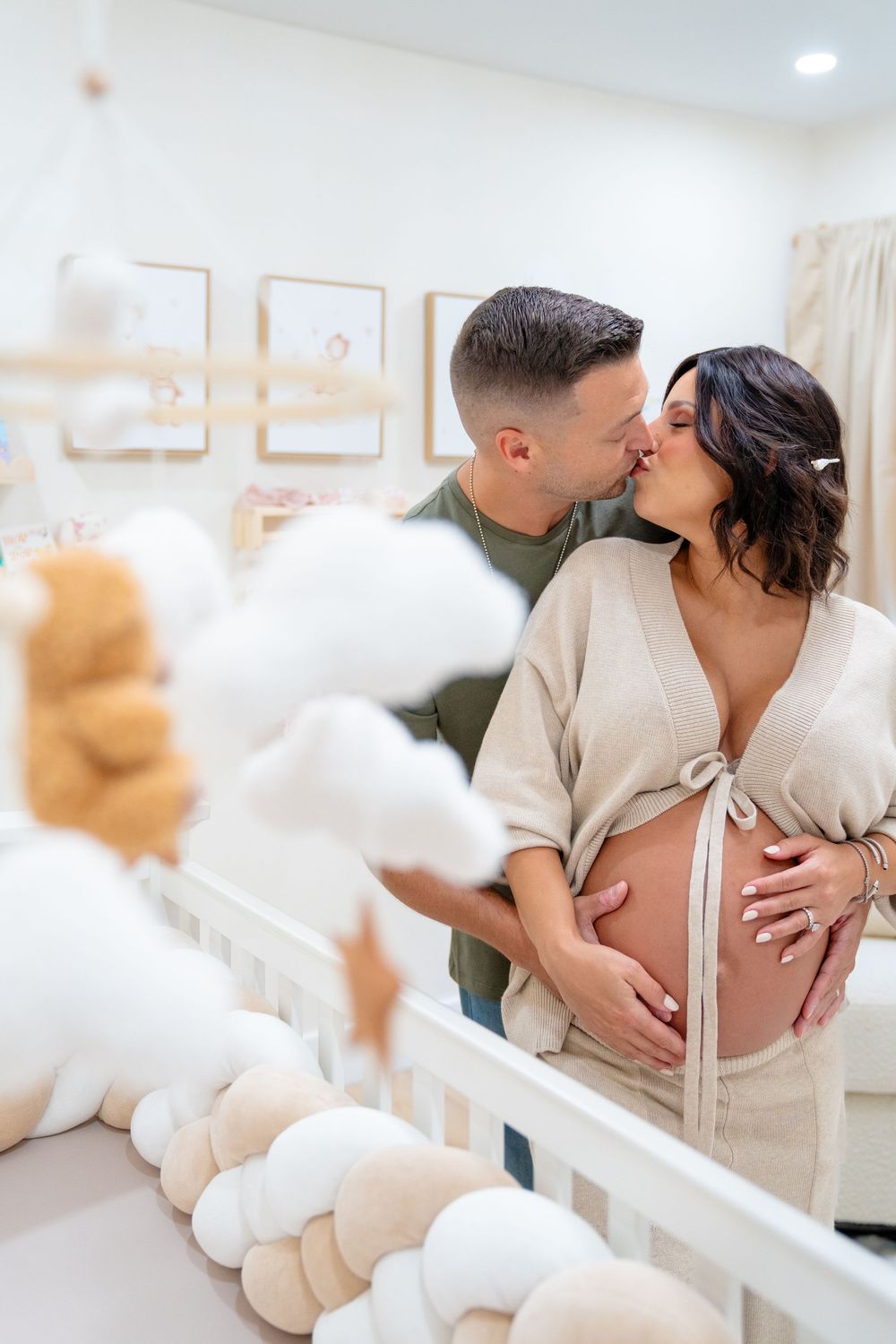 A romantic maternity photo shows a couple sharing an intimate moment in a nursery with soft white decor and teddy bears.