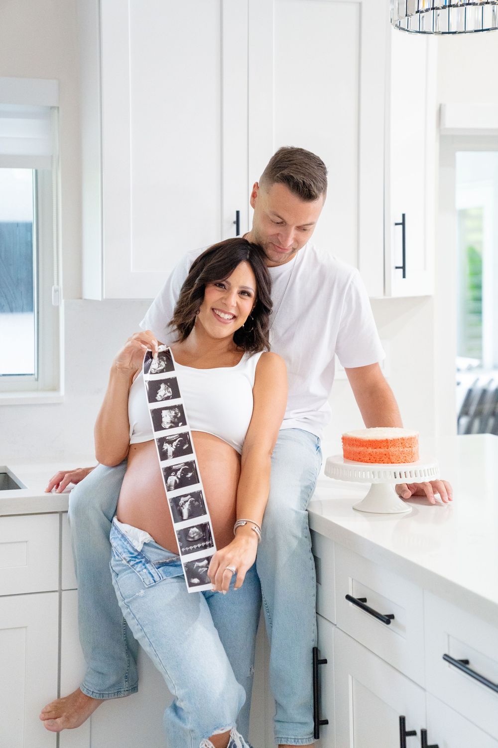 Expectant couple poses in white kitchen while holding ultrasound photos.