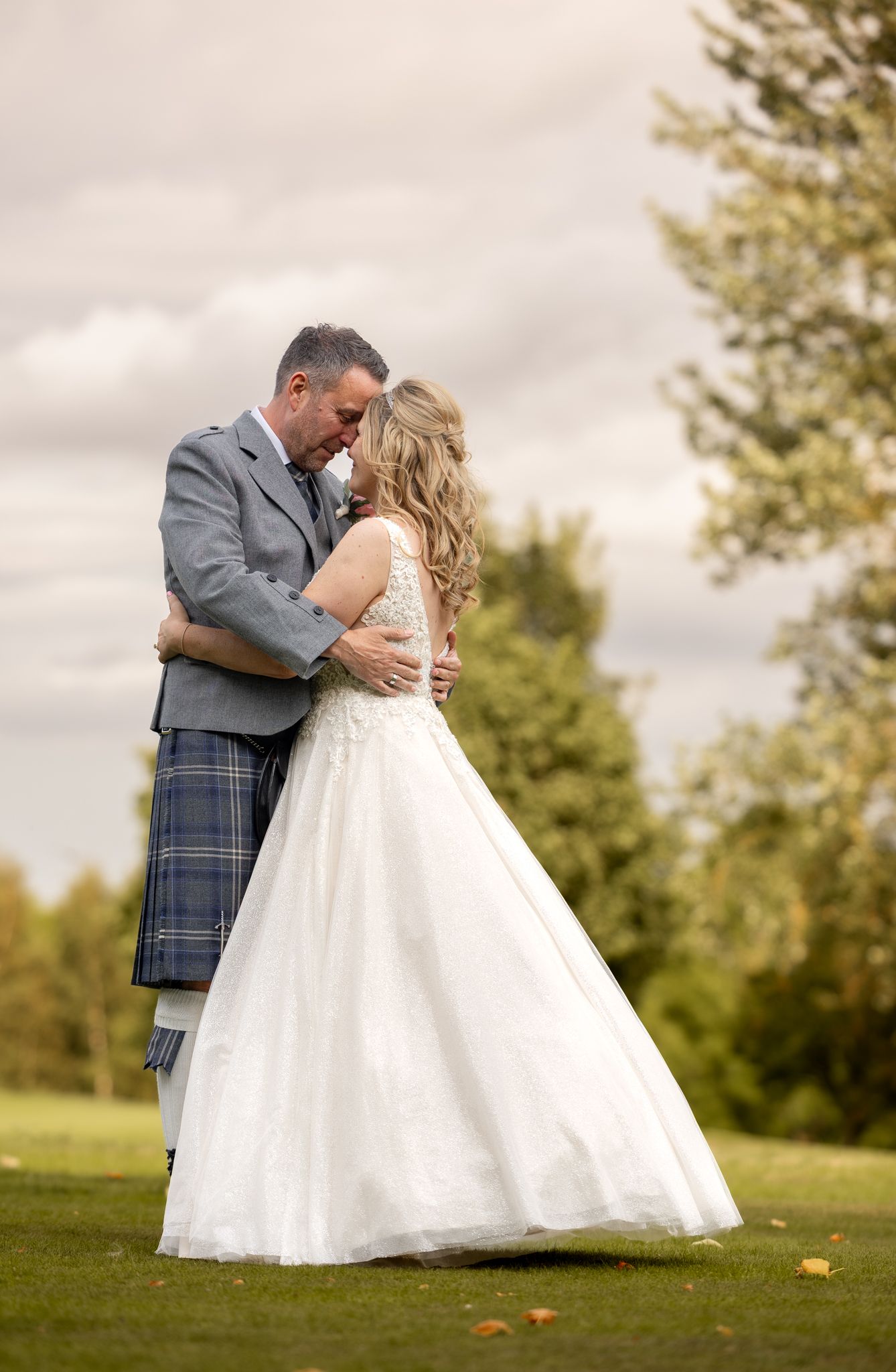 A couple shares a romantic moment on their wedding day, with the groom wearing a traditional Scottish kilt on a grassy hill.