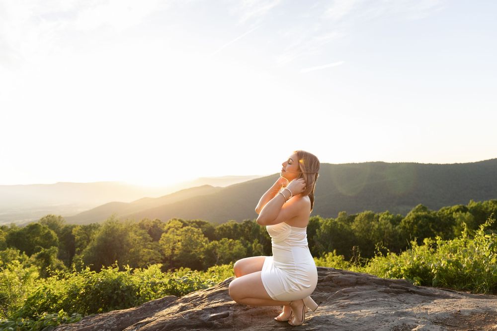 Woman in white dress meditating on rock overlooking lush green valley at sunset.