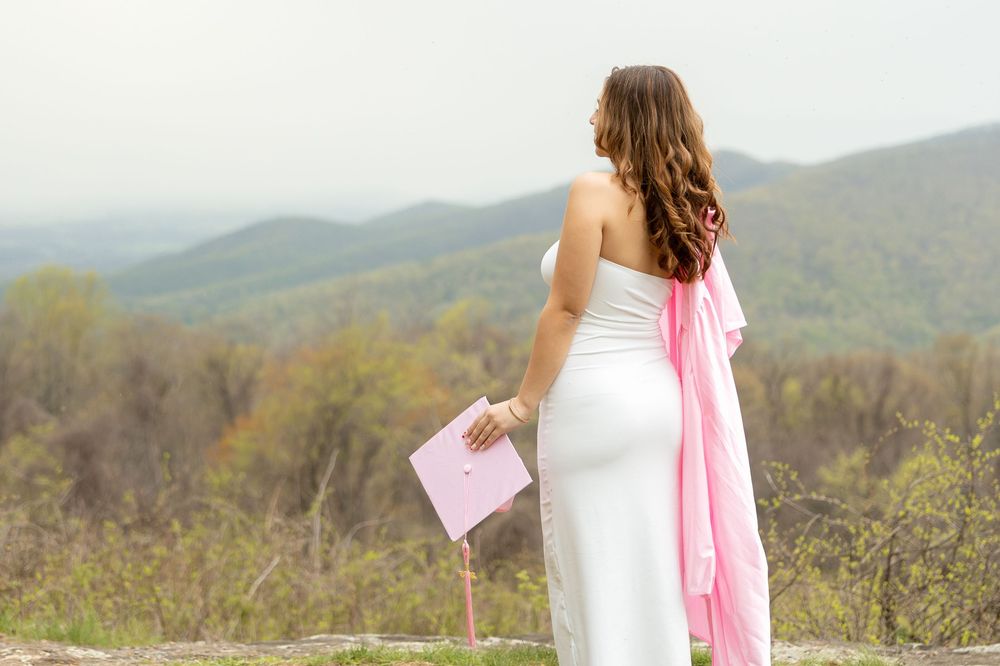 Figure in white dress with pink accessories stands on mountain overlook holding envelope against scenic mountain backdrop.