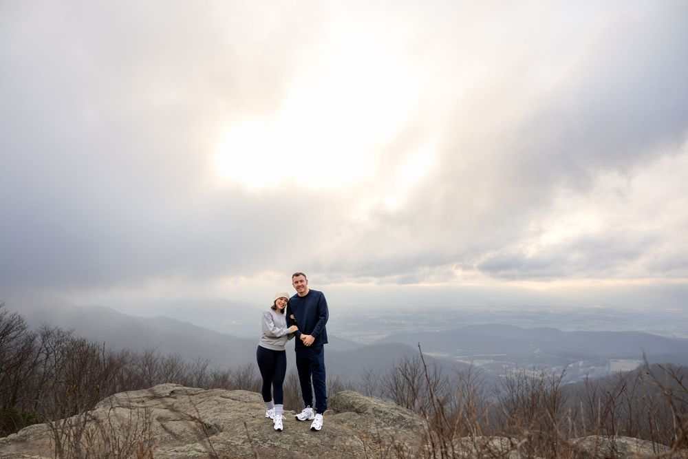 A couple stands together at the summit of a mountain overlook with cloudy skies and misty valleys stretching into the distance.