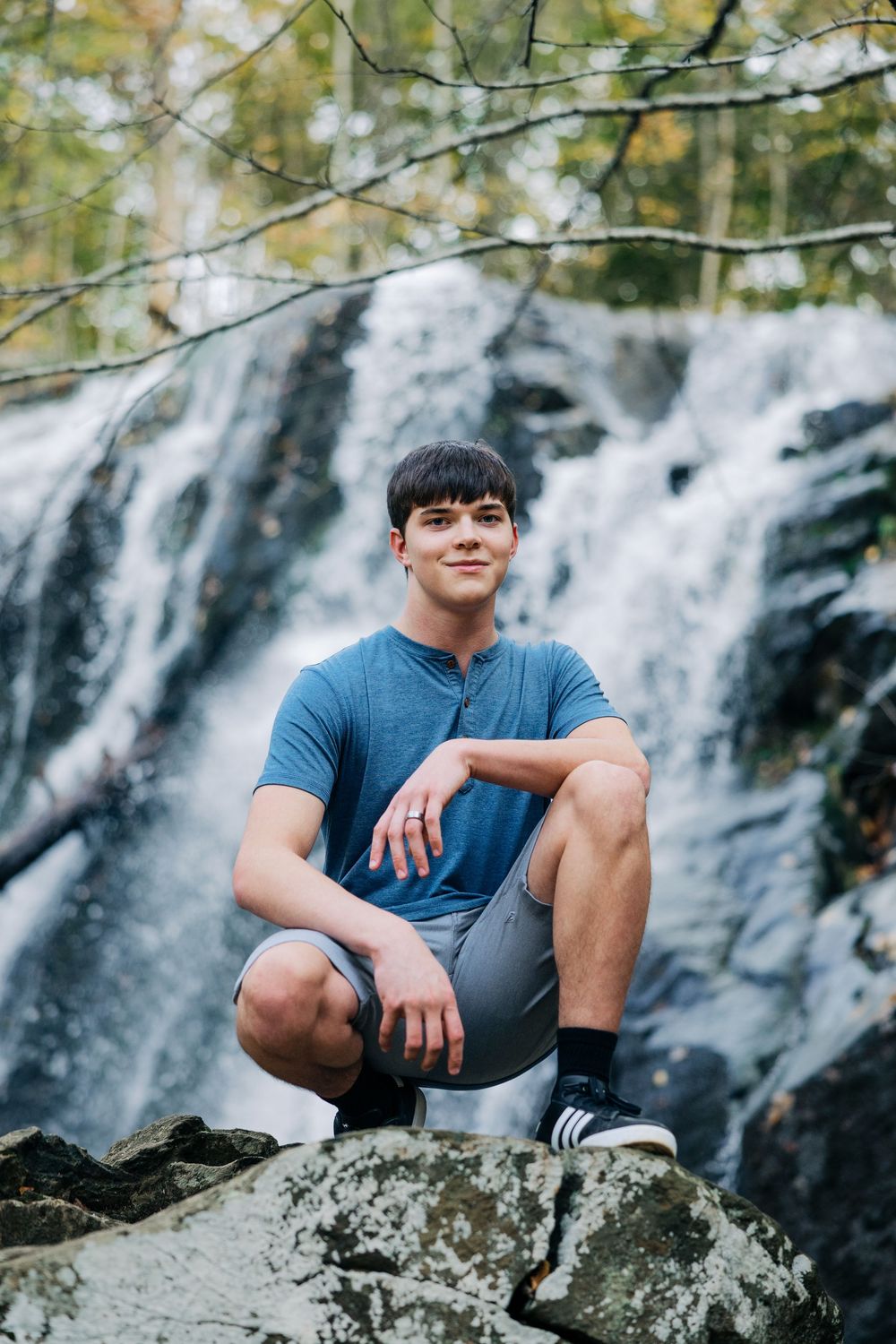 A figure crouches on a rocky outcrop against a cascading waterfall backdrop while wearing a blue t-shirt and athletic shorts.