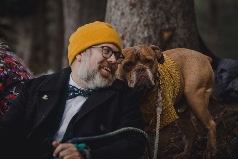 Brown dog in yellow scarf joins companion wearing yellow beanie on forest trail.