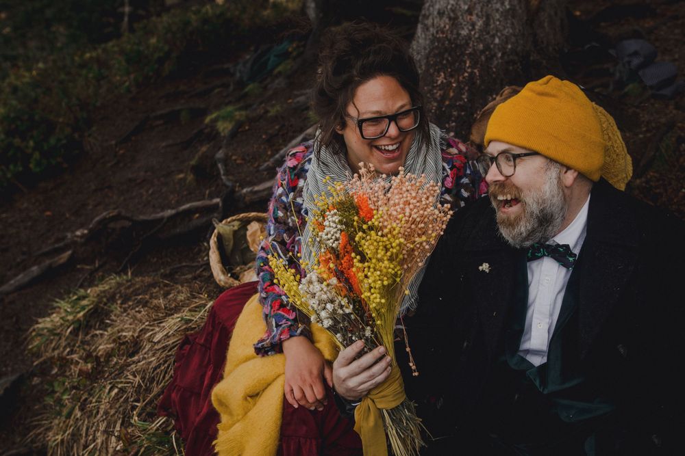 Couple laughs while holding vibrant wildflower bouquet in forest setting.