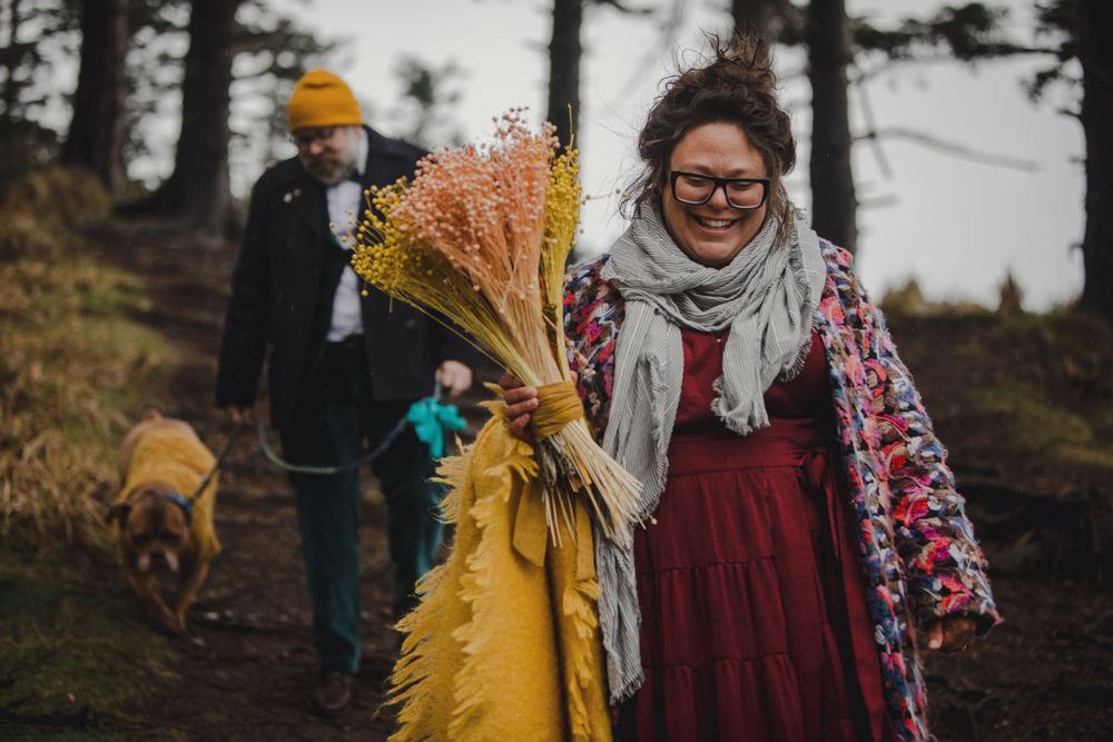 Two people and a dog walk along a misty forest trail with dried flowers and autumn colors.