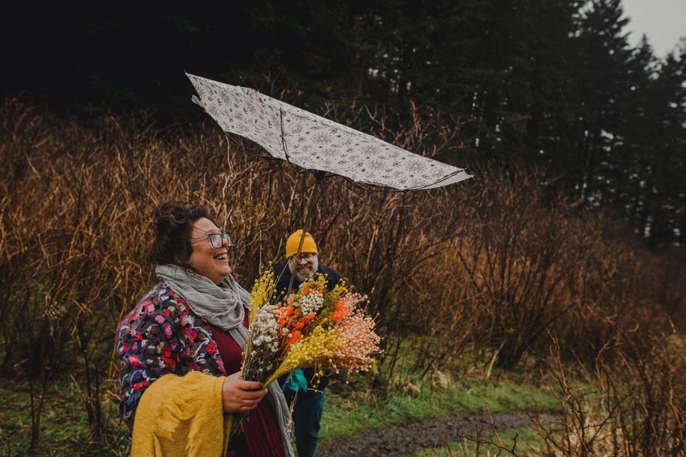 A couple shares a joyful moment under a white umbrella holding colorful flowers on a misty hillside.