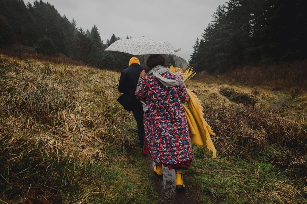 A person in a colorful coat and yellow boots holds up a white umbrella on a grassy hillside.