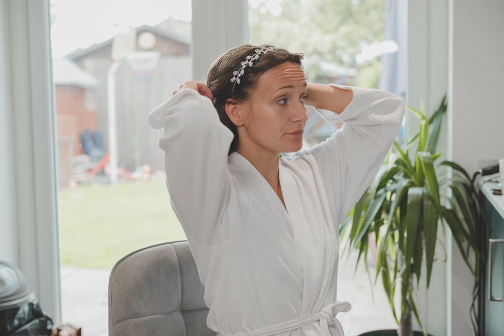 Woman in white robe adjusting hair accessory near bright window.
