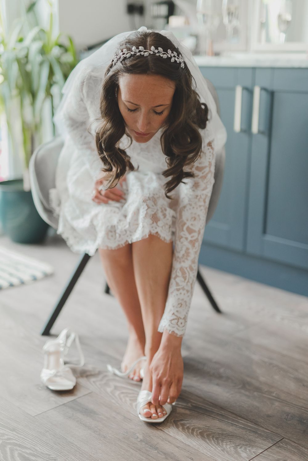 A bride in lace wedding dress bends down to put on white sandals before her ceremony.