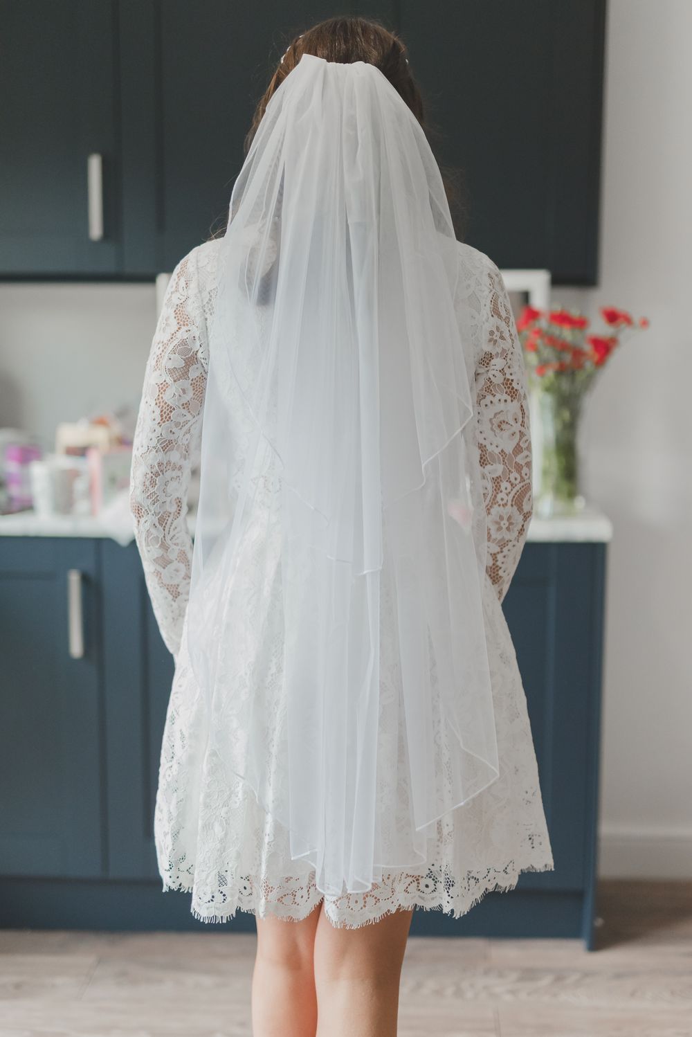Back view of a short lace wedding dress with flowing veil against blue kitchen cabinets.