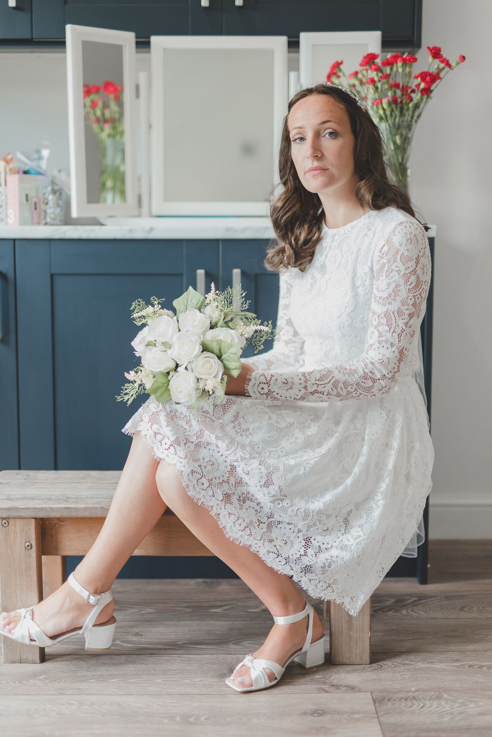 Bride in short lace dress holds white bouquet while sitting on wooden bench in modern kitchen.