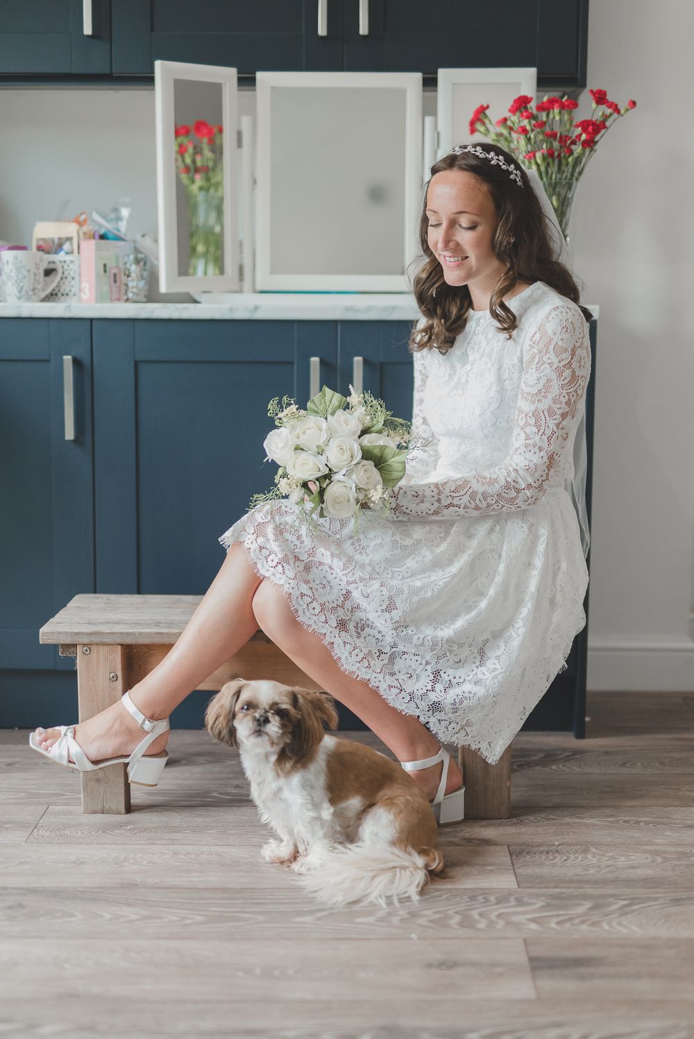 Bride in lace dress sits with white bouquet as small dog poses beside her on wooden bench.