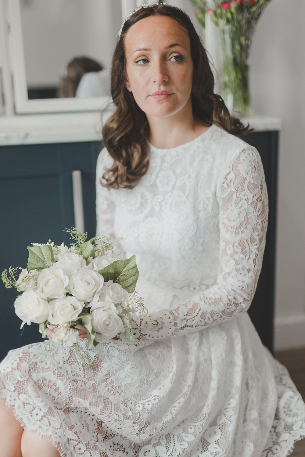 Elegant bridal portrait featuring white lace dress and classic white rose bouquet against blue cabinetry.
