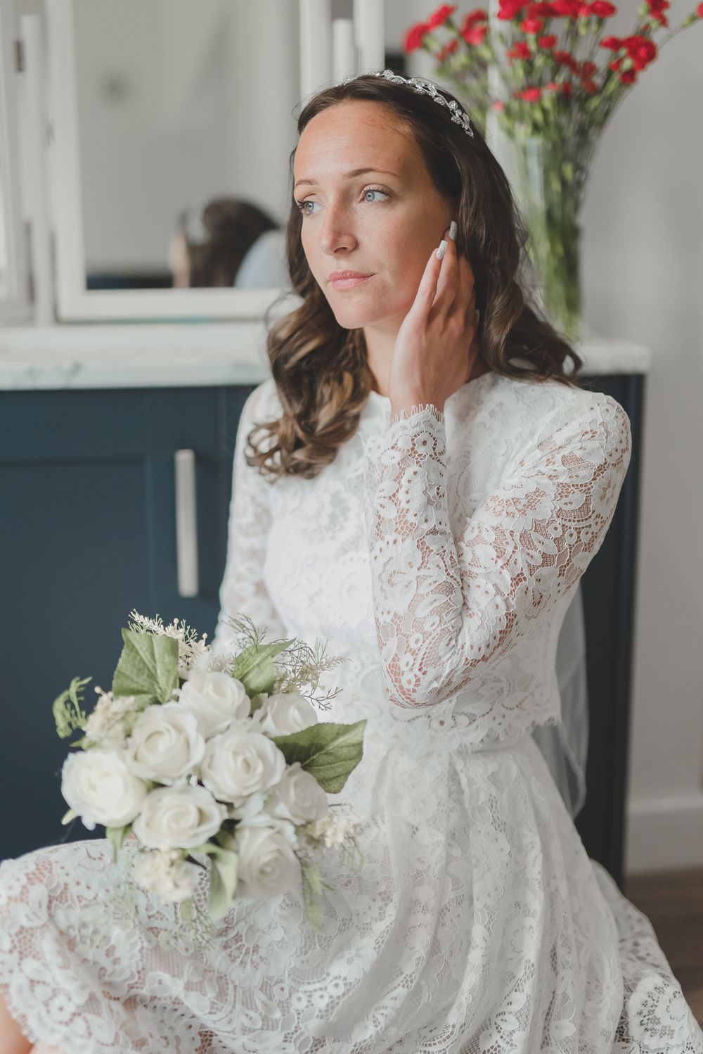 Bride in white lace dress with wavy hair holds white rose bouquet while touching her face near red flowers in vase.