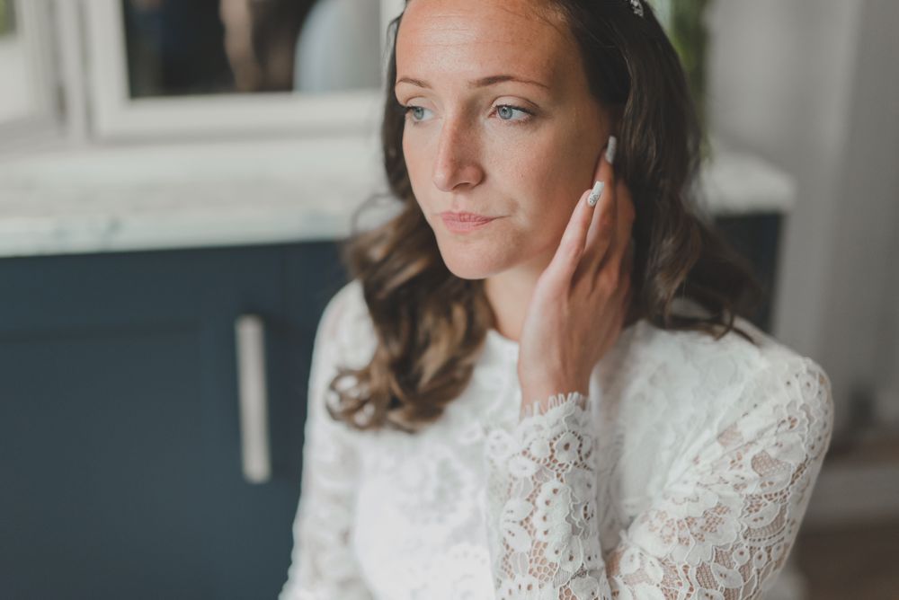Close up portrait of bride in lace dress with wavy hair and natural makeup touching her cheek.