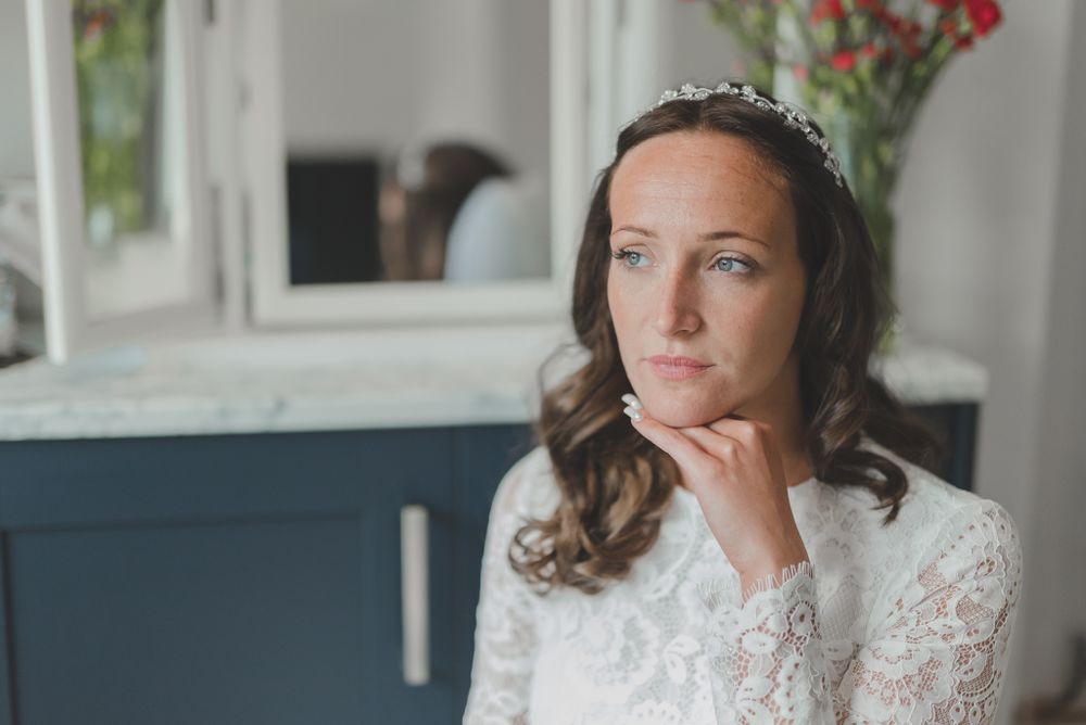 Thoughtful bride in lace dress and tiara poses with hand under chin near window and red flowers.