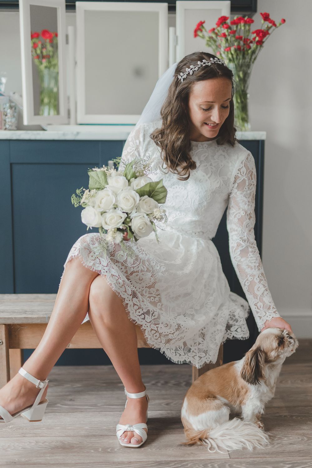 Smiling bride in short lace dress sits with small dog while holding white rose bouquet.