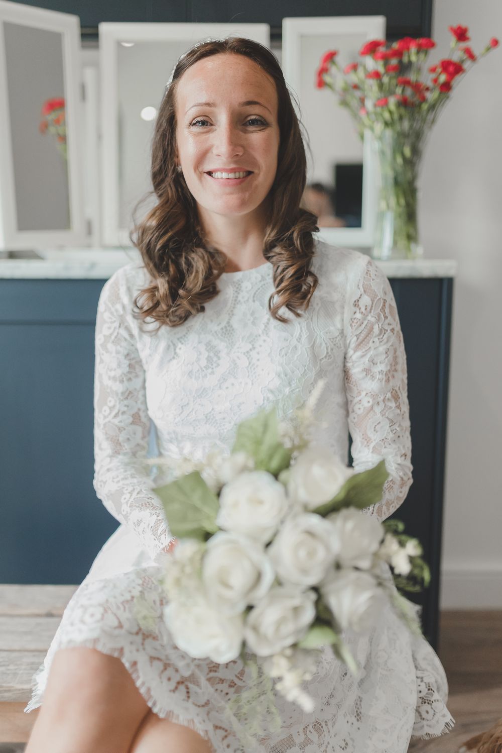 Joyful bride in short lace wedding dress holds white rose bouquet while sitting.