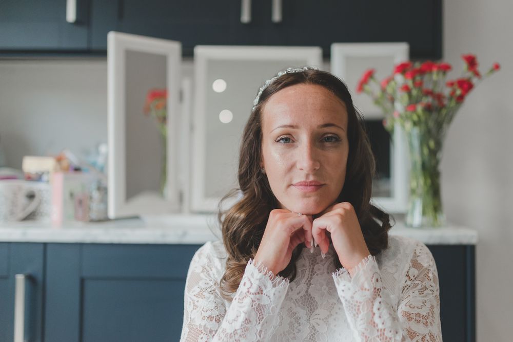 Portrait of bride in lace dress resting chin on hands with navy cabinets and red flowers in background.