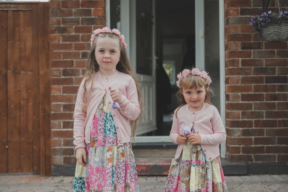 Two young flower girls in floral dresses and pink cardigans stand outside brick building.