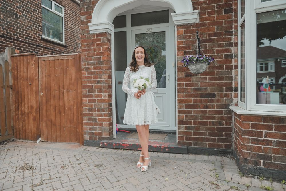 Bride in short white dress poses in doorway of brick house holding bouquet.