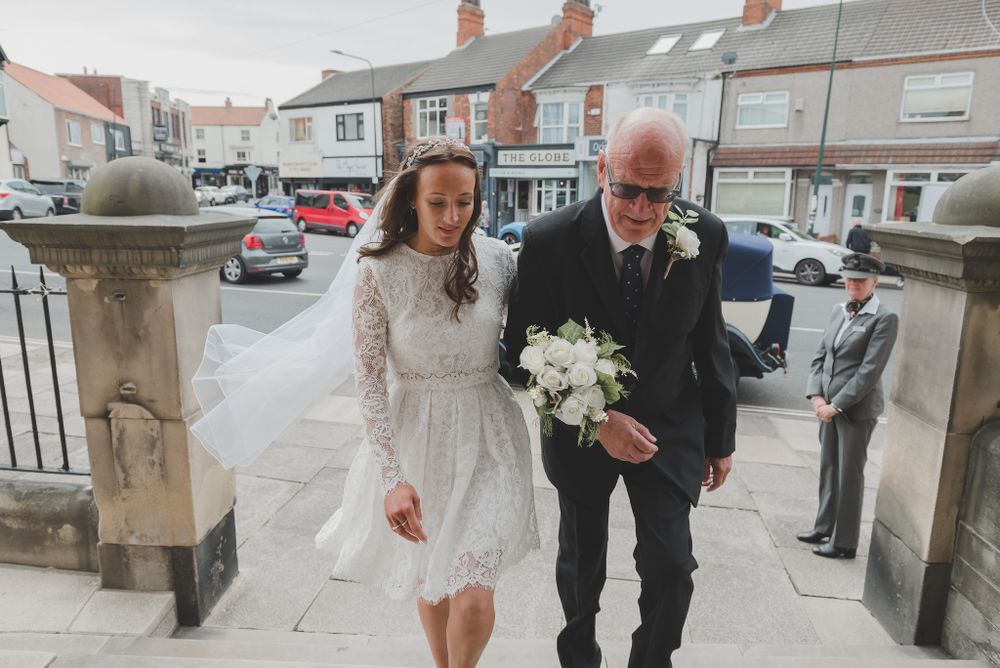 A bride in a short lace dress walks up stone steps with her escort.