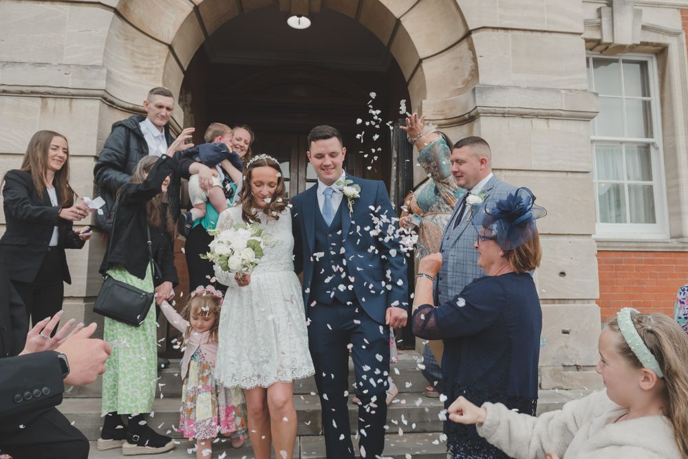 Wedding party celebrates with confetti throw outside historic building entrance.