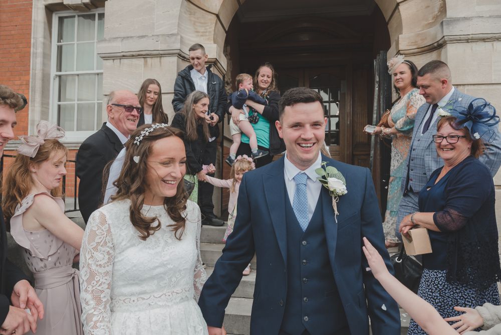 Newlyweds exit stone building surrounded by joyful guests throwing confetti on their wedding day.