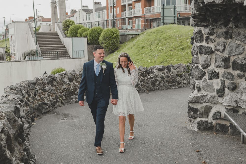 Couple walks along stone wall path with modern buildings in background.