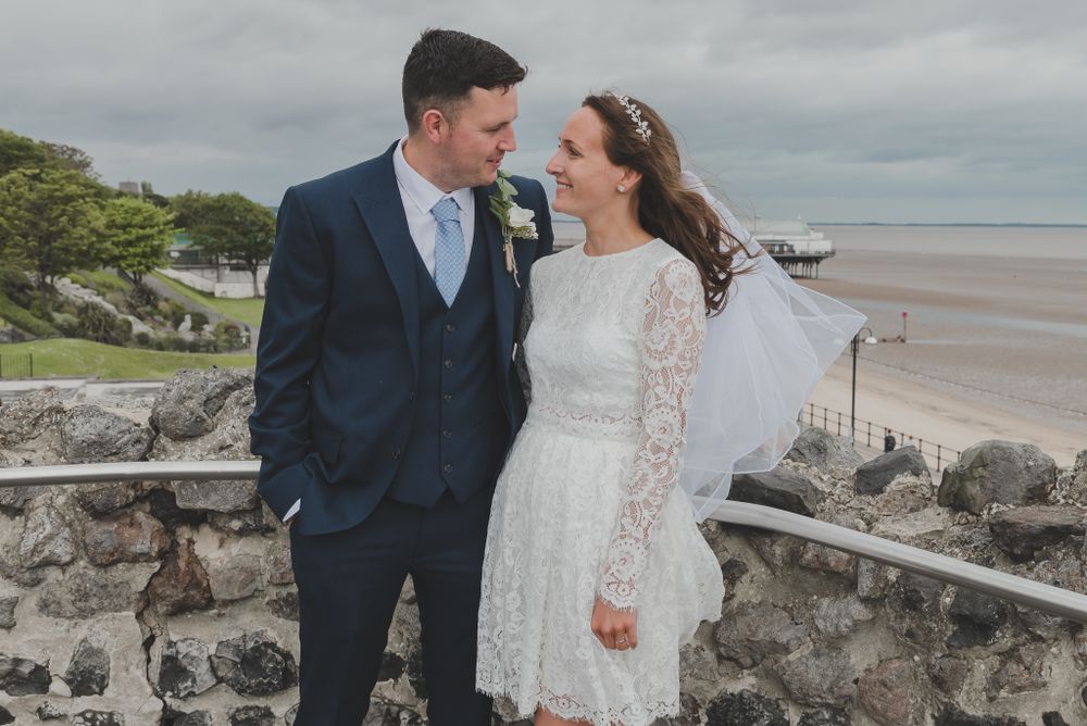Coastal wedding portrait with dramatic cloudy sky and stone wall overlooking beach.