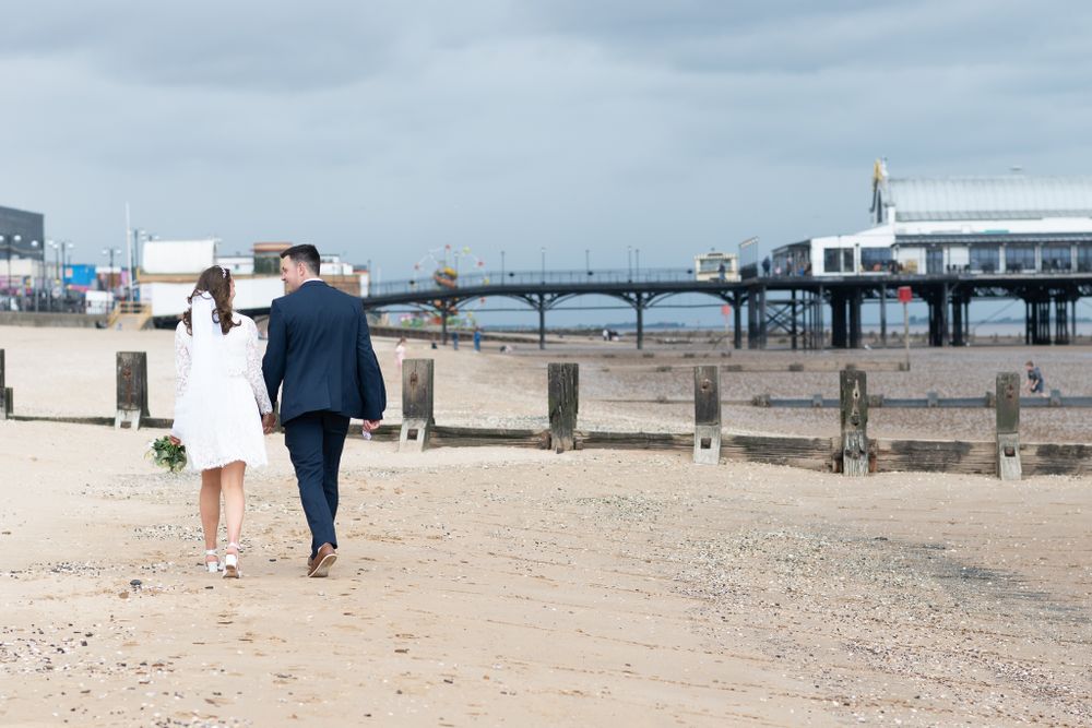 A couple walking away on a beach boardwalk with a pier and building visible in the background.