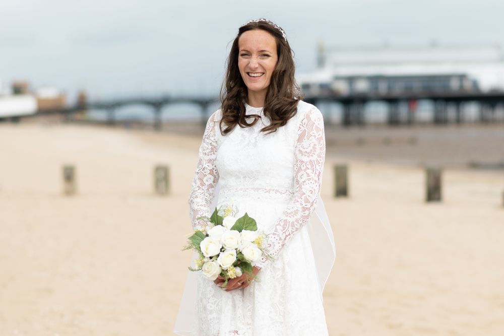 Bride in white lace dress holding bouquet stands on beach with pier in background.