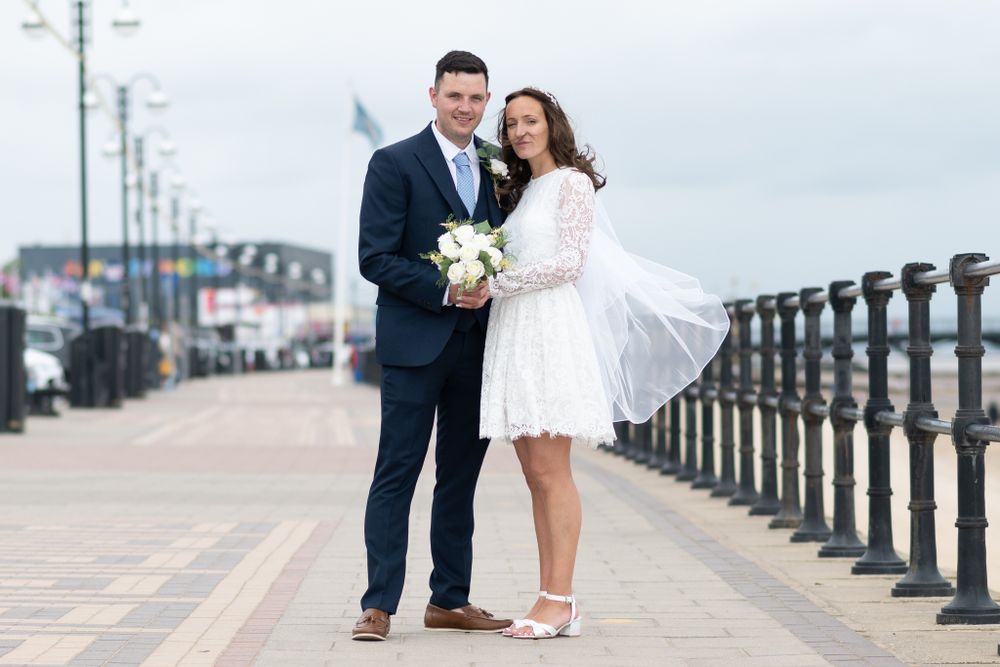 Wedding couple poses along seaside promenade with historic pier in background.