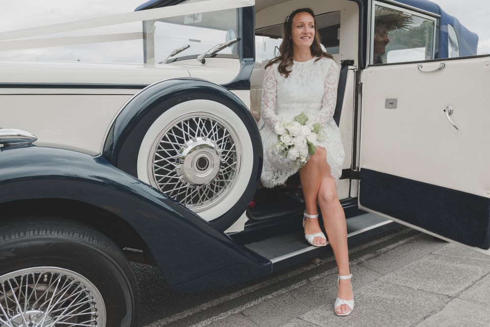 Bride in vintage-style dress poses elegantly by classic car.