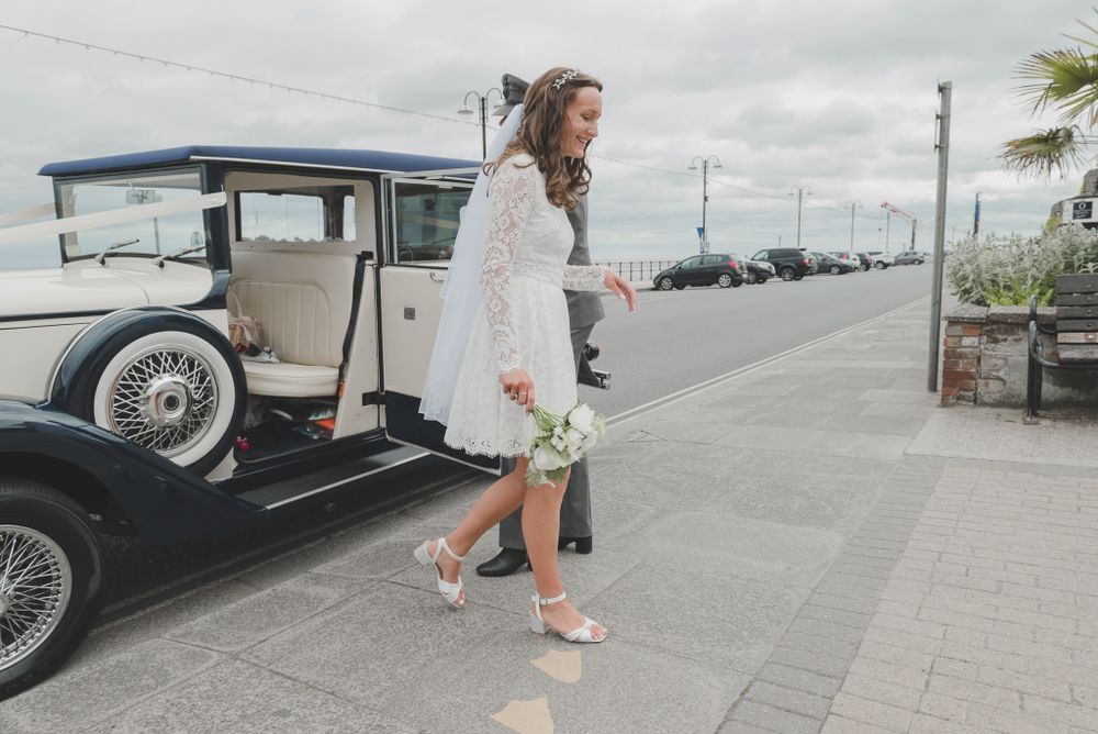 A bride in a white dress steps out of a vintage car on a cloudy day by the seaside.
