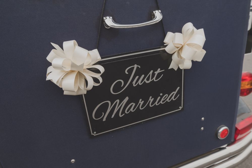 A 'Just Married' sign decorated with white flowers hangs on a dark car.