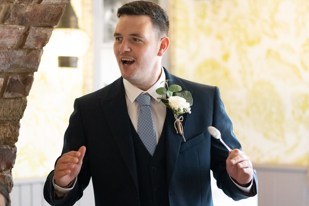 A groom in navy suit and grey tie gestures expressively during a speech.