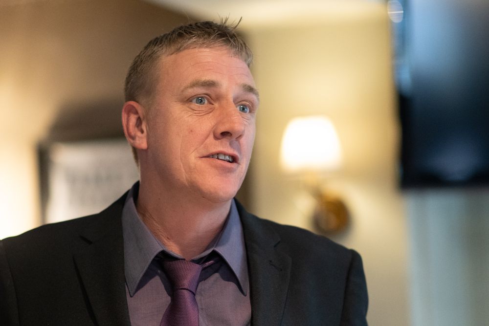 A person in formal attire speaks with an expressive gesture against a warm-lit background.