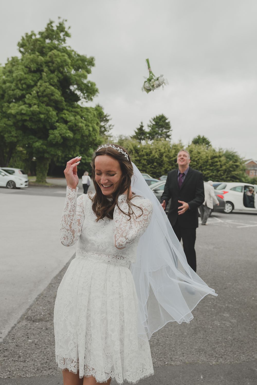 Bride in lace dress celebrates throwing her bouquet in parking lot.