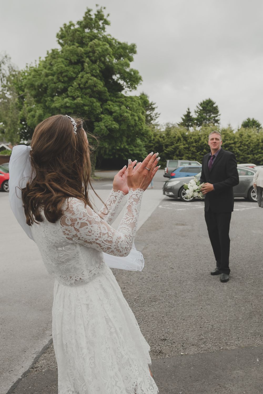 Bride in white lace gown reaches for tossed bouquet in outdoor setting.